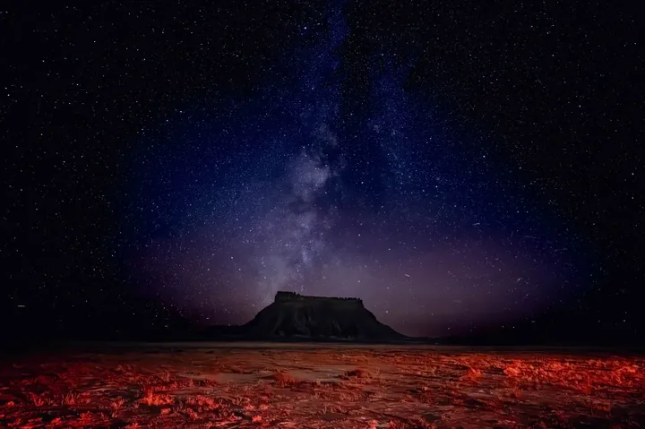 Factory Butte is silhouetted against a cosmic sea of stars, as the desert burns crimson below.